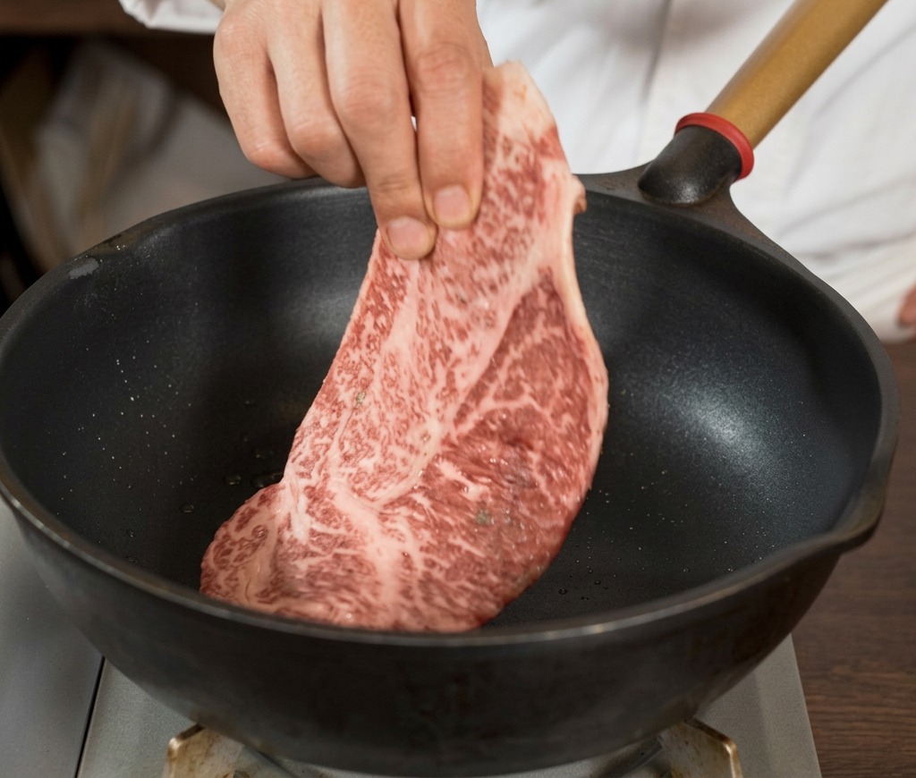 Una mano colocando un filete crudo, grueso y veteado, sazonado con pimienta negra, en una sartén negra bien caliente.