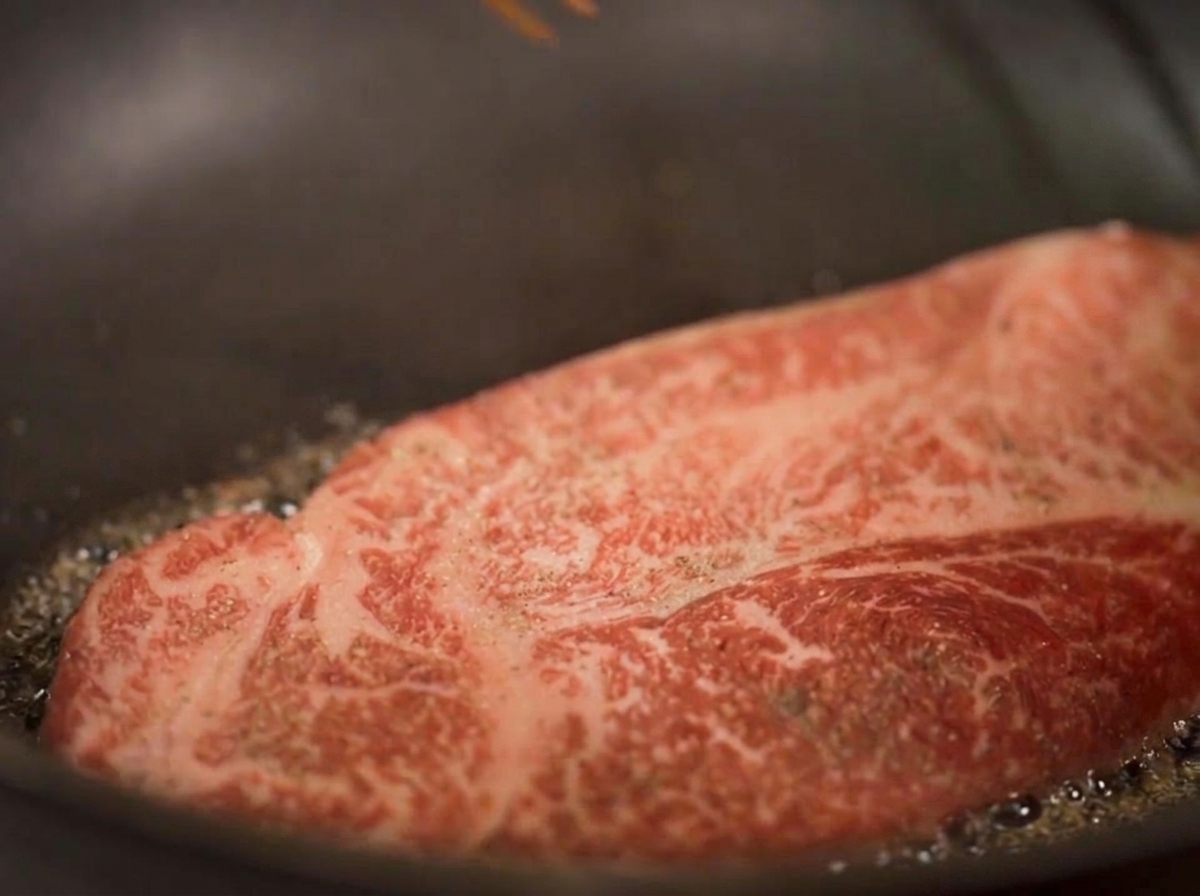 Primer plano de un filete grueso sellándose en aceite, con vapor visible y los bordes inferiores tornándose de un color marrón dorado.