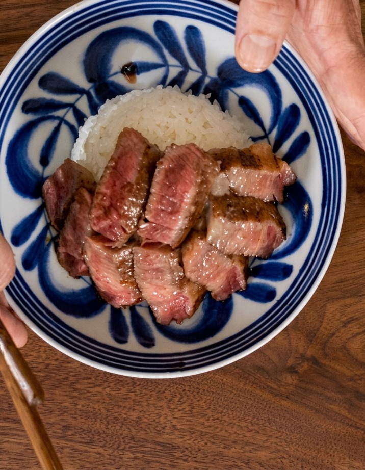 Vista cenital de una mano sosteniendo un bol con detalles azules mientras las láminas de filete se colocan en abanico sobre una cama de arroz blanco.