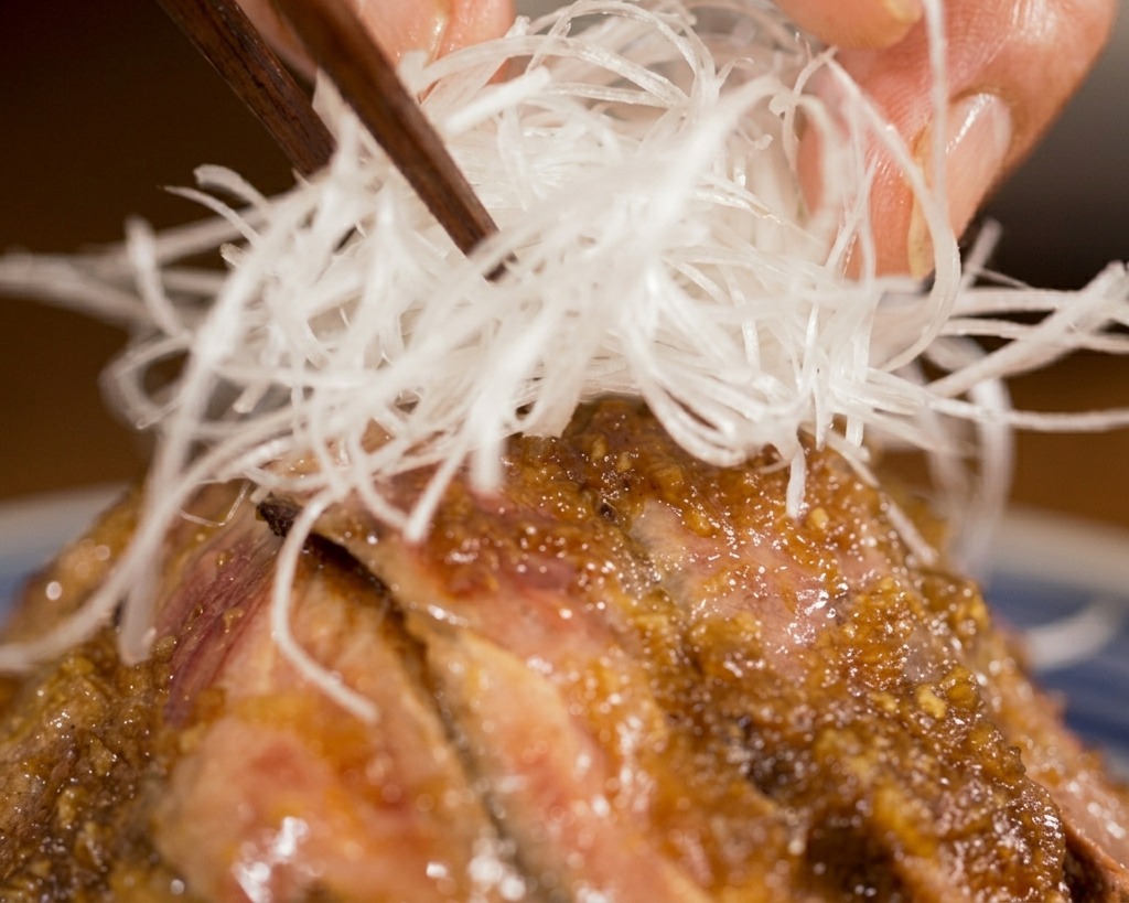 Palillos colocando un nido de finas tiras blancas de puerro en el centro de un bol de steak donburi con salsa.