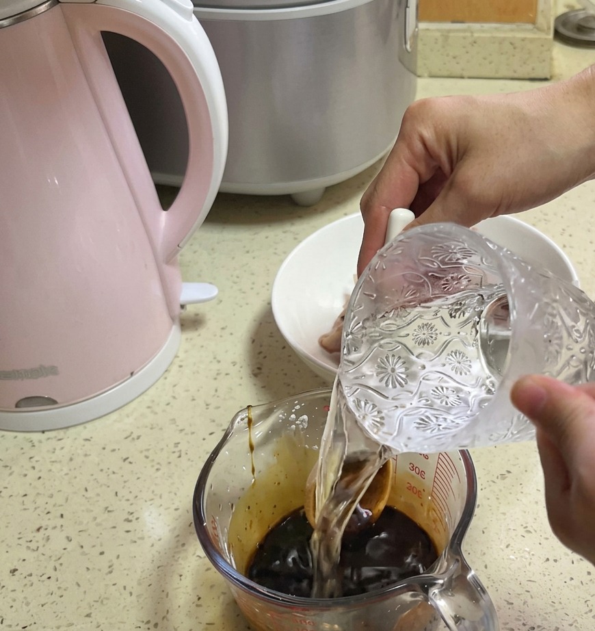 Agua limpia vertiéndose desde una jarra de cristal decorativa en una taza medidora que contiene una base de salsa de color marrón oscuro.
