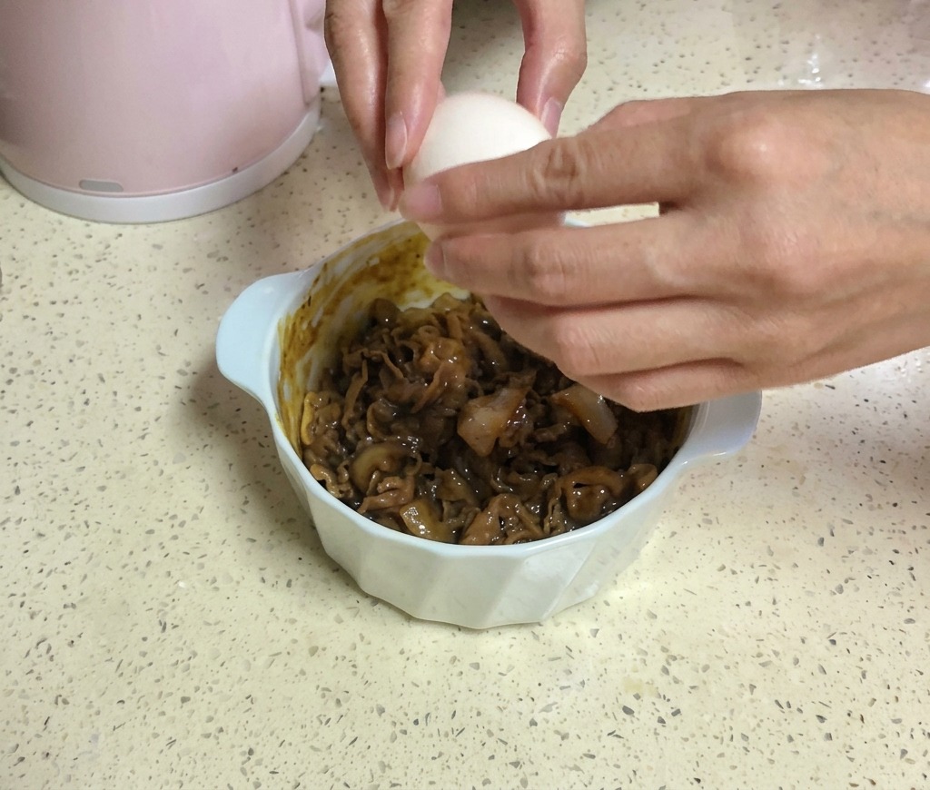 Dos manos cascando un huevo blanco sobre un tazón de ternera cubierto con dos lonchas de queso amarillo.