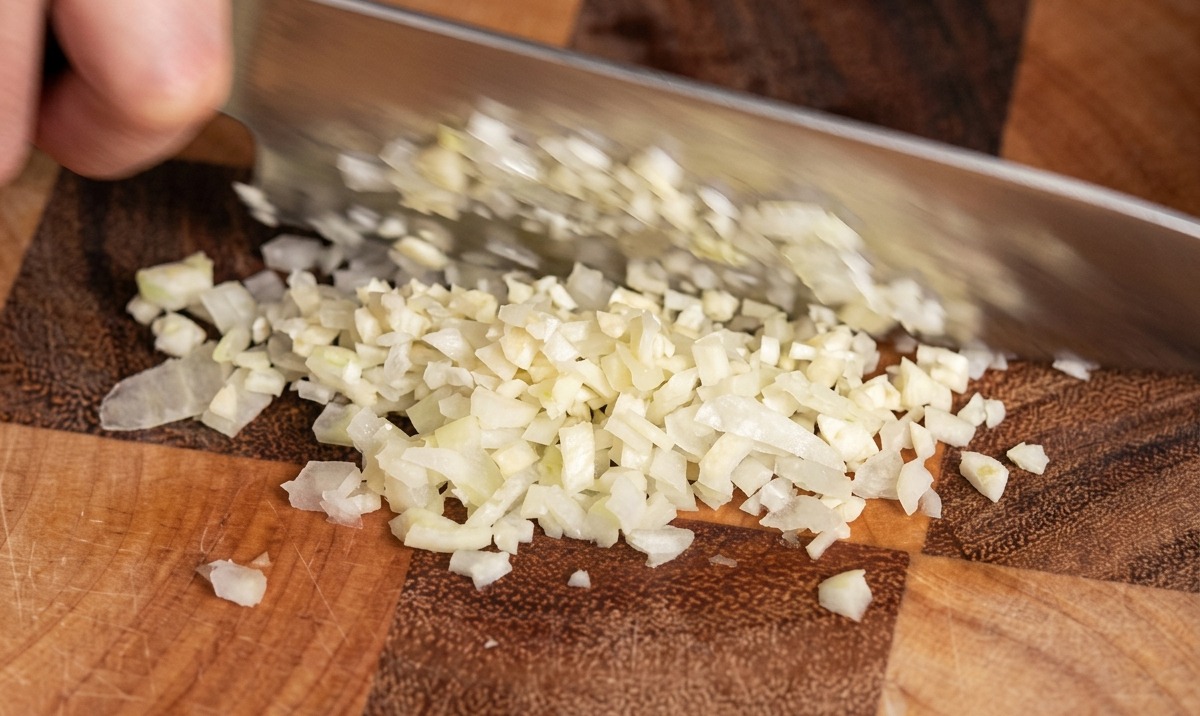 Un primer plano del cuchillo de un chef picando dientes de ajo en trozos pequeños sobre una tabla de cortar de madera.