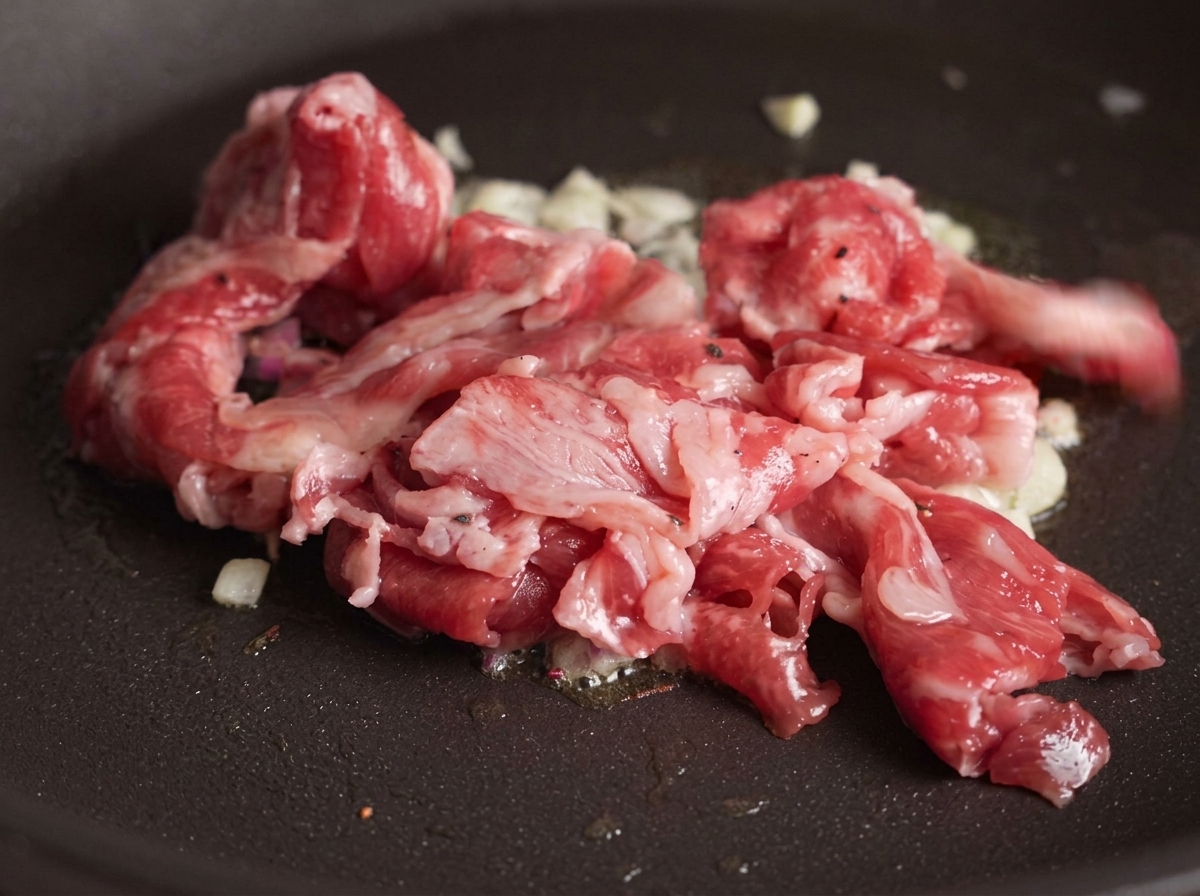 Láminas de ternera cruda añadiéndose a una sartén con ajo salteado, con algunas partes de la carne empezando a dorarse por el calor.