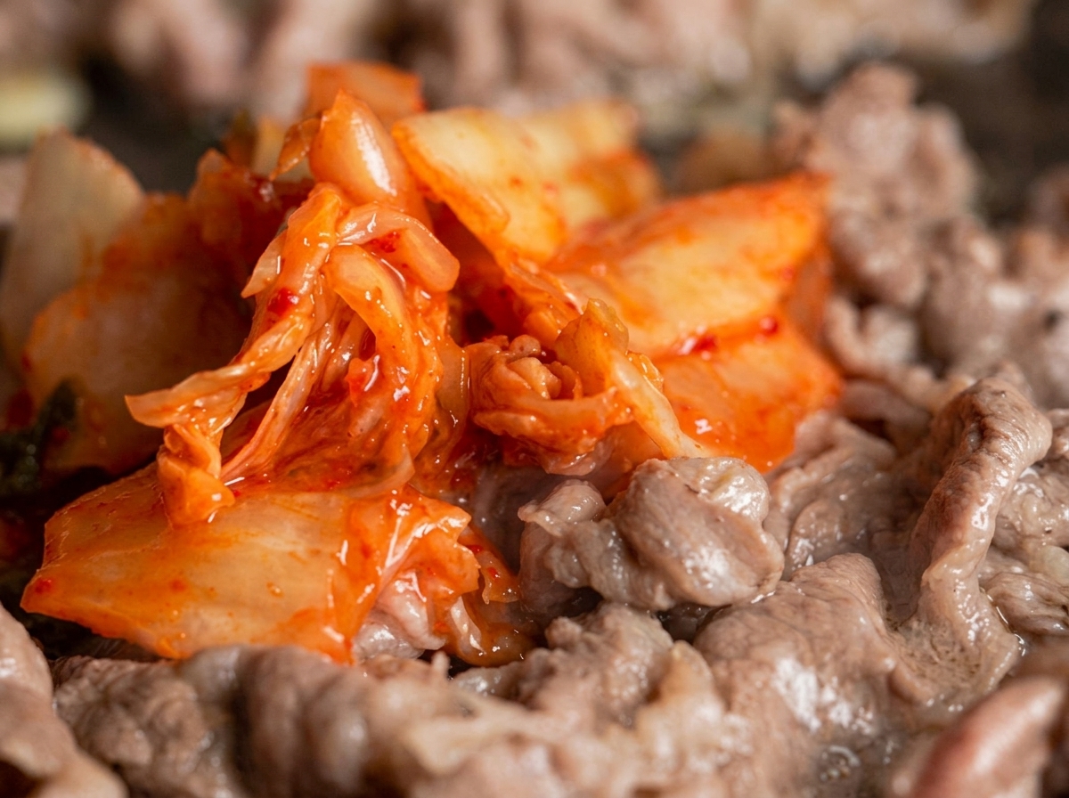 Una porción generosa de kimchi de color rojo brillante mezclándose con tiras de ternera finamente cortadas en una sartén.