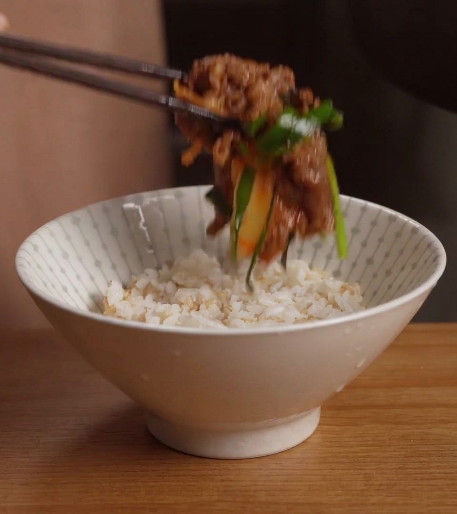 Unos palillos colocando una porción de ternera salteada con kimchi en un bol blanco lleno de arroz cocido.