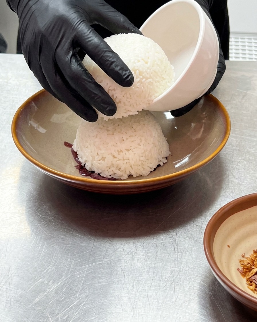 Manos con guantes levantando un tazón blanco pequeño para revelar un domo de arroz blanco al vapor sobre la cebolla morada.