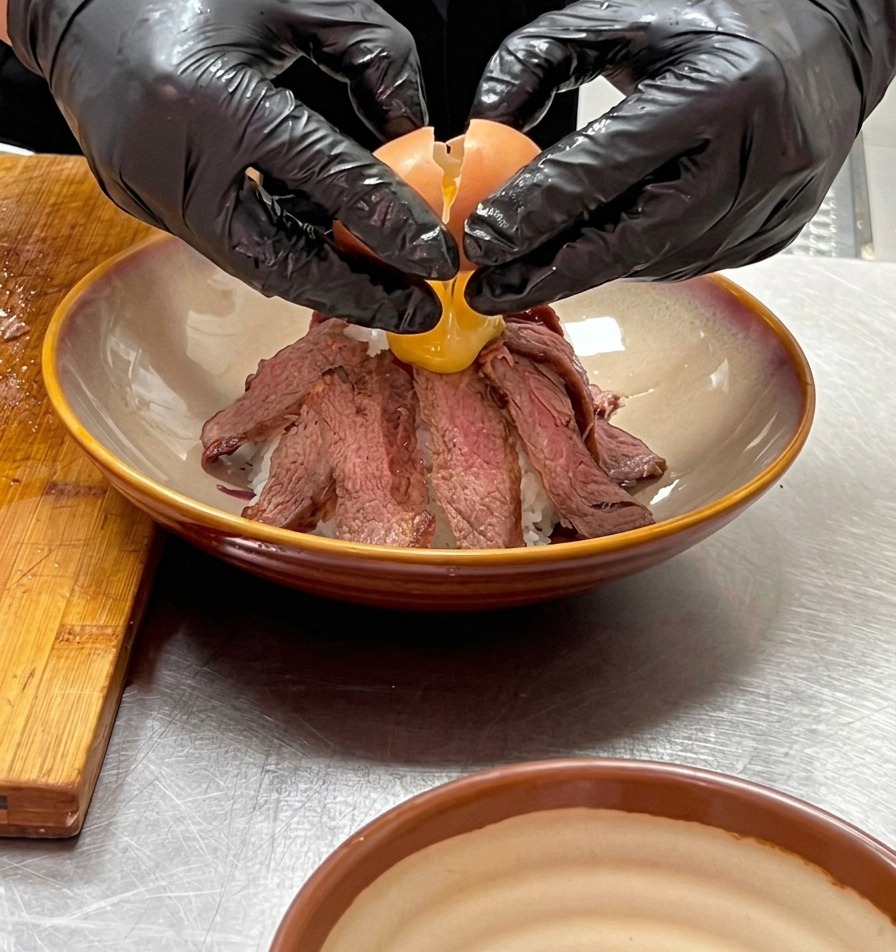 Dos manos con guantes cascando con cuidado un huevo onsen sobre la cima de la montaña de carne troceada.