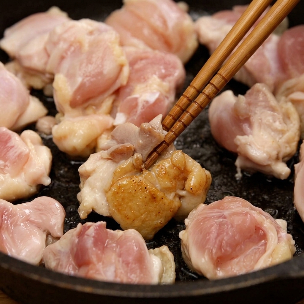 Trozos de contramuslo de pollo salteándose en una sartén con palillos de madera hasta que se doran ligeramente.
