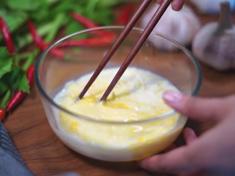 Un par de palillos de madera oscuros batiendo una mezcla de huevo, leche y queso dentro de un tazón de vidrio.