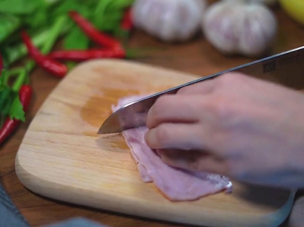 Primer plano de unas manos usando un cuchillo de acero inoxidable para cortar tiras de tocino crudo en cubos pequeños sobre una tabla de madera.