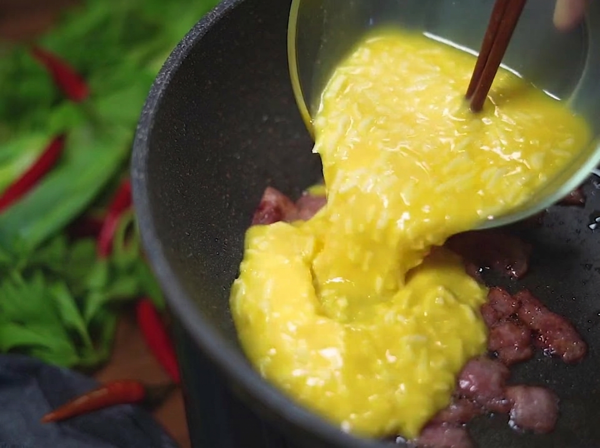 Una mezcla amarilla de huevo y queso vertiéndose desde un tazón de vidrio en una sartén caliente con trozos de tocino crujiente.