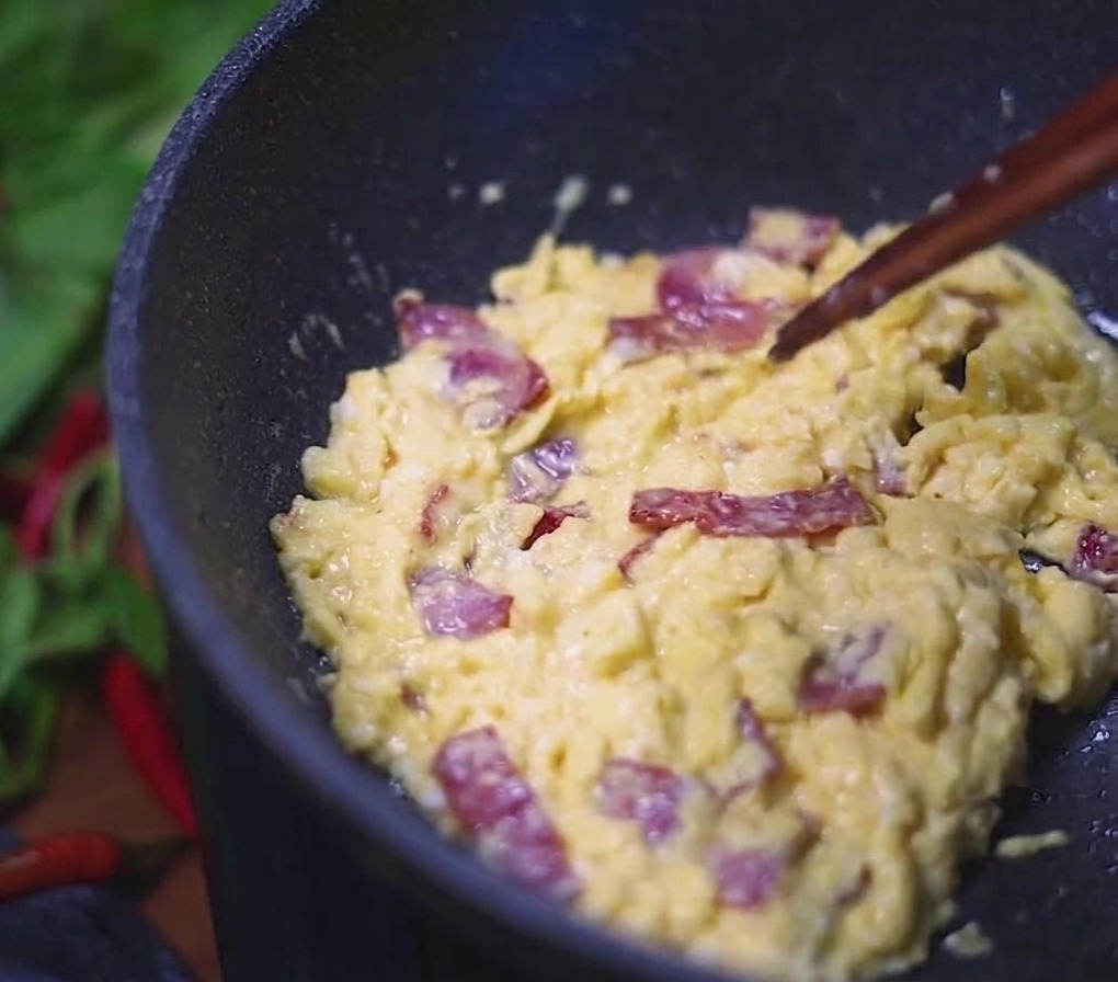 Huevos revueltos cremosos mezclados con trozos de tocino revolviéndose con palillos en una sartén oscura.