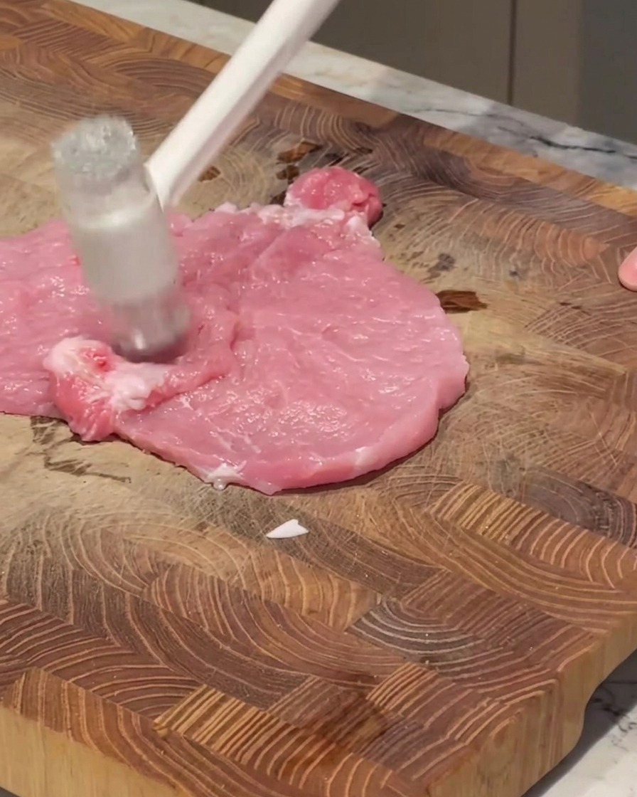 Un mazo de cocina blanco golpeando un filete de cerdo crudo sobre una tabla de madera.