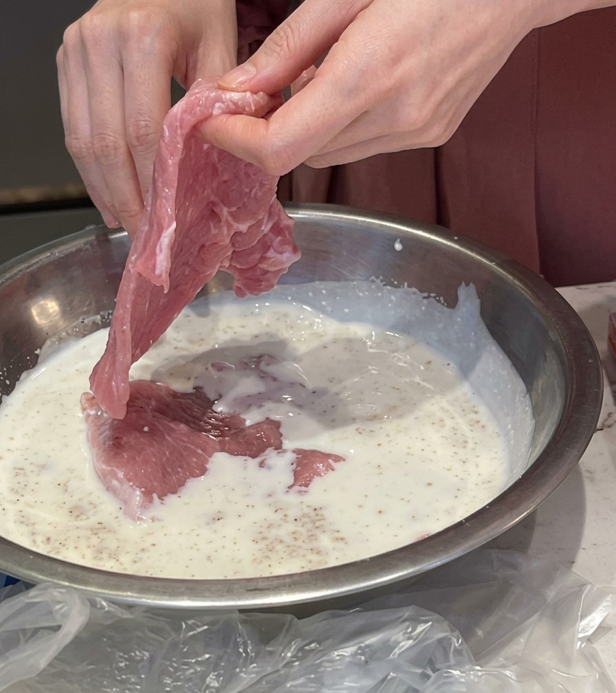 Manos sumergiendo un filete de cerdo rosado en un tazón metálico lleno de leche con ajo.