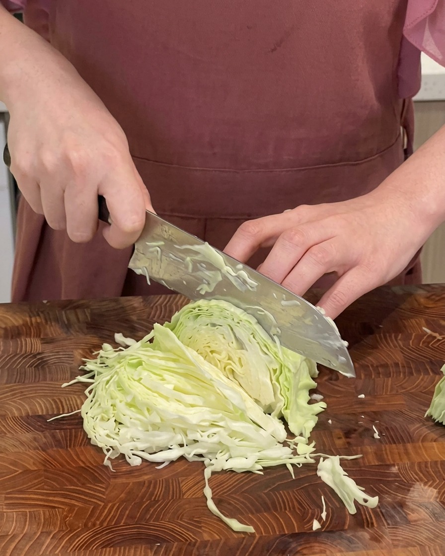 Un chef picando finamente un repollo verde en tiras delgadas sobre una tabla de cortar con un cuchillo grande.