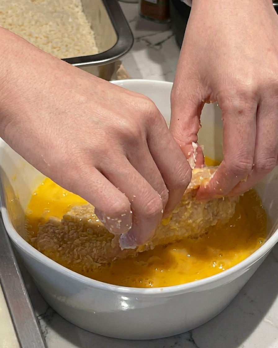 Una mano sumergiendo un filete de cerdo enharinado en un tazón de cerámica blanca con huevo batido.