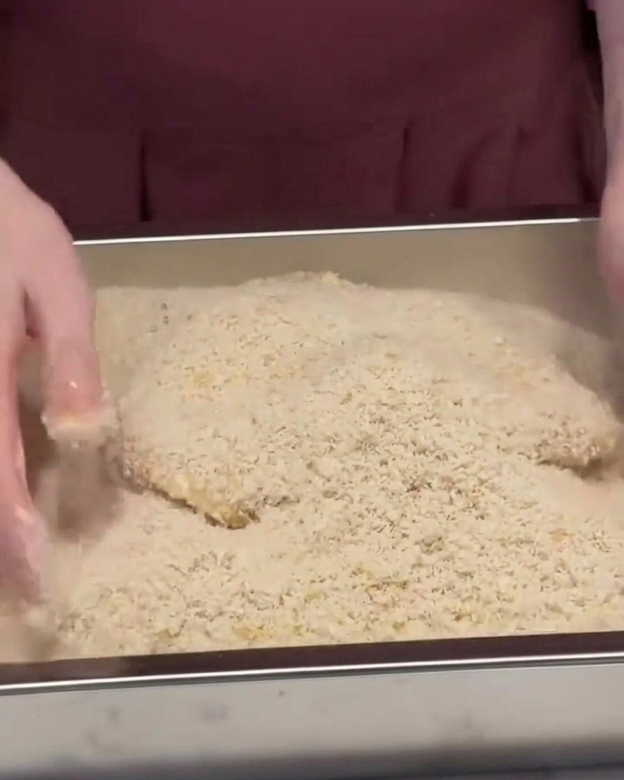 Un filete de cerdo siendo cubierto y presionado en una bandeja metálica llena de pan rallado panko.