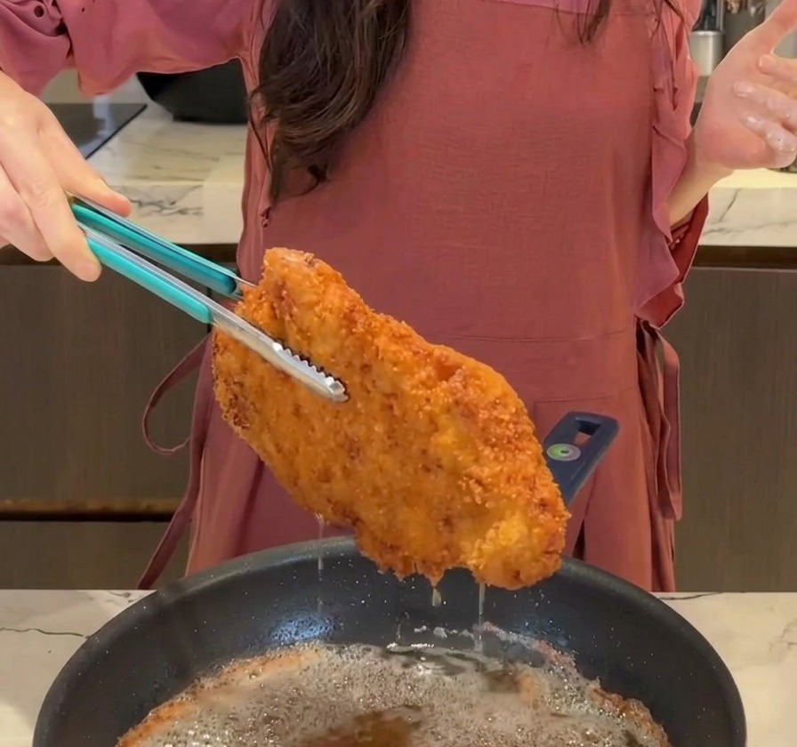 Una chuleta de cerdo frita, dorada y crujiente siendo retirada de la sartén con unas pinzas azules mientras escurre el aceite.