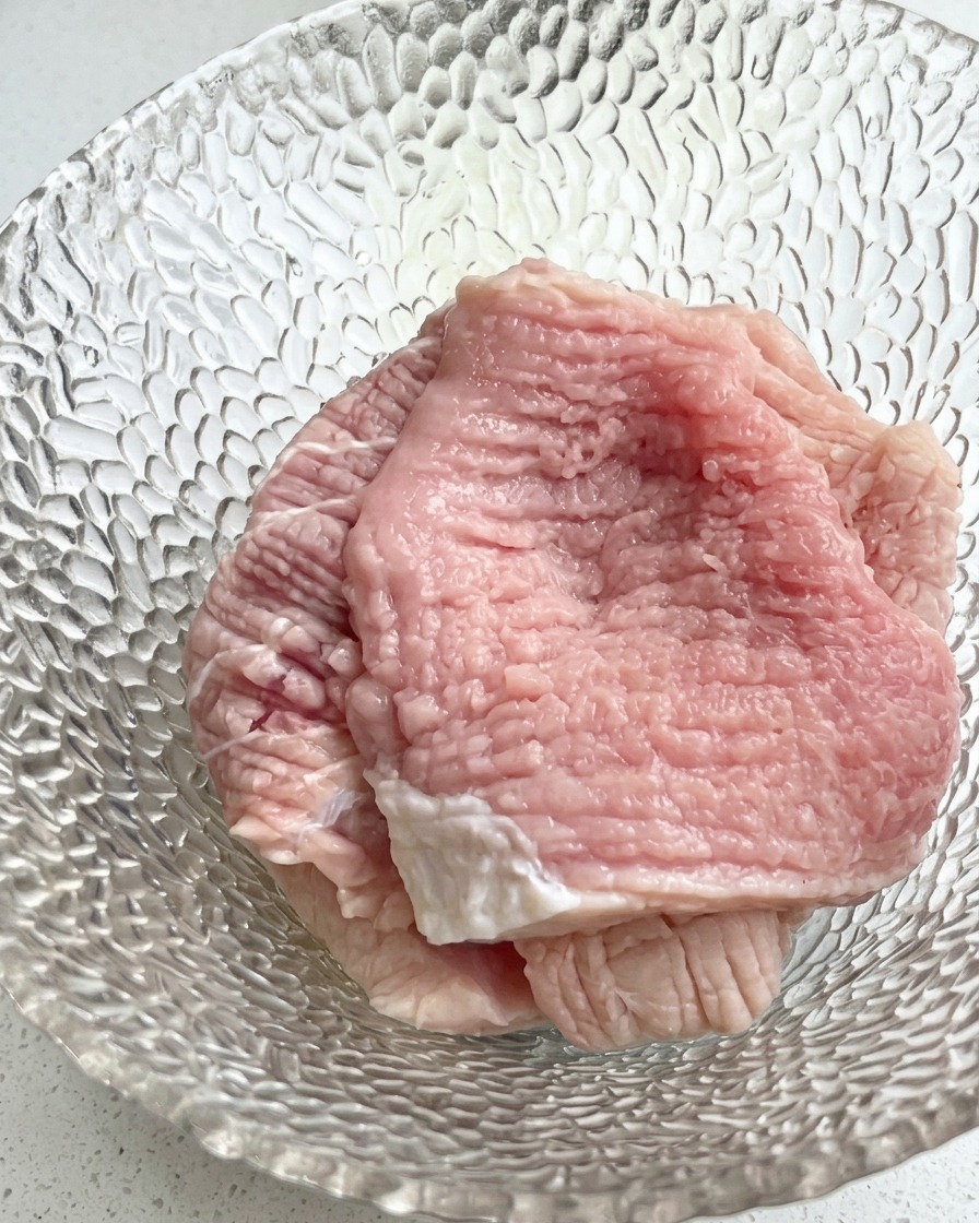 Vista de cerca de un bol de vidrio con filetes de cerdo crudos cubiertos con cebolleta picada y condimentos líquidos.