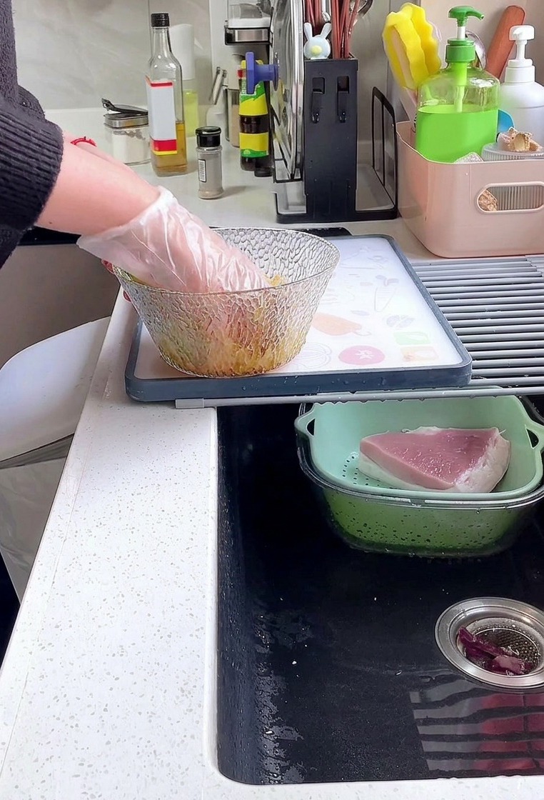 Una persona con guante de plástico mezcla filetes de lomo de cerdo con salsa de soja, pimienta y almidón en un bol de vidrio.