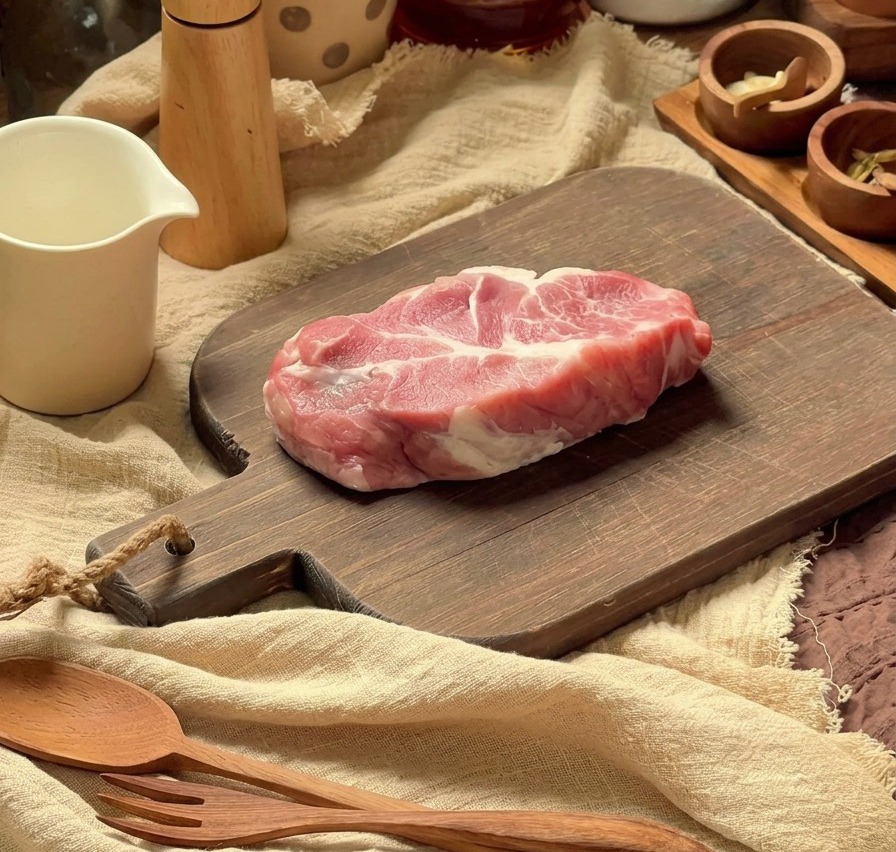 Chuleta de cerdo cruda preparándose en una tabla de cortar de madera con utensilios de cocina al fondo.