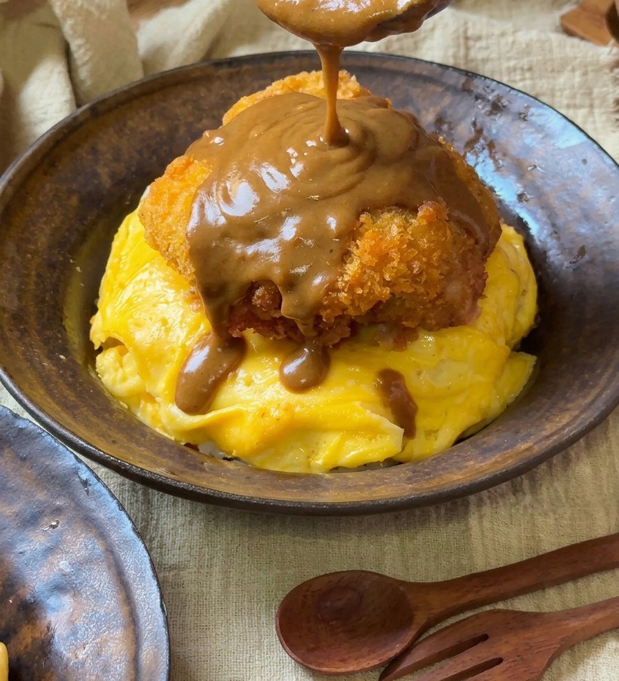 Una salsa de curry espesa y marrón vertiéndose desde una cuchara de madera sobre una chuleta de cerdo dorada que descansa sobre una cama de huevos amarillos en espiral.