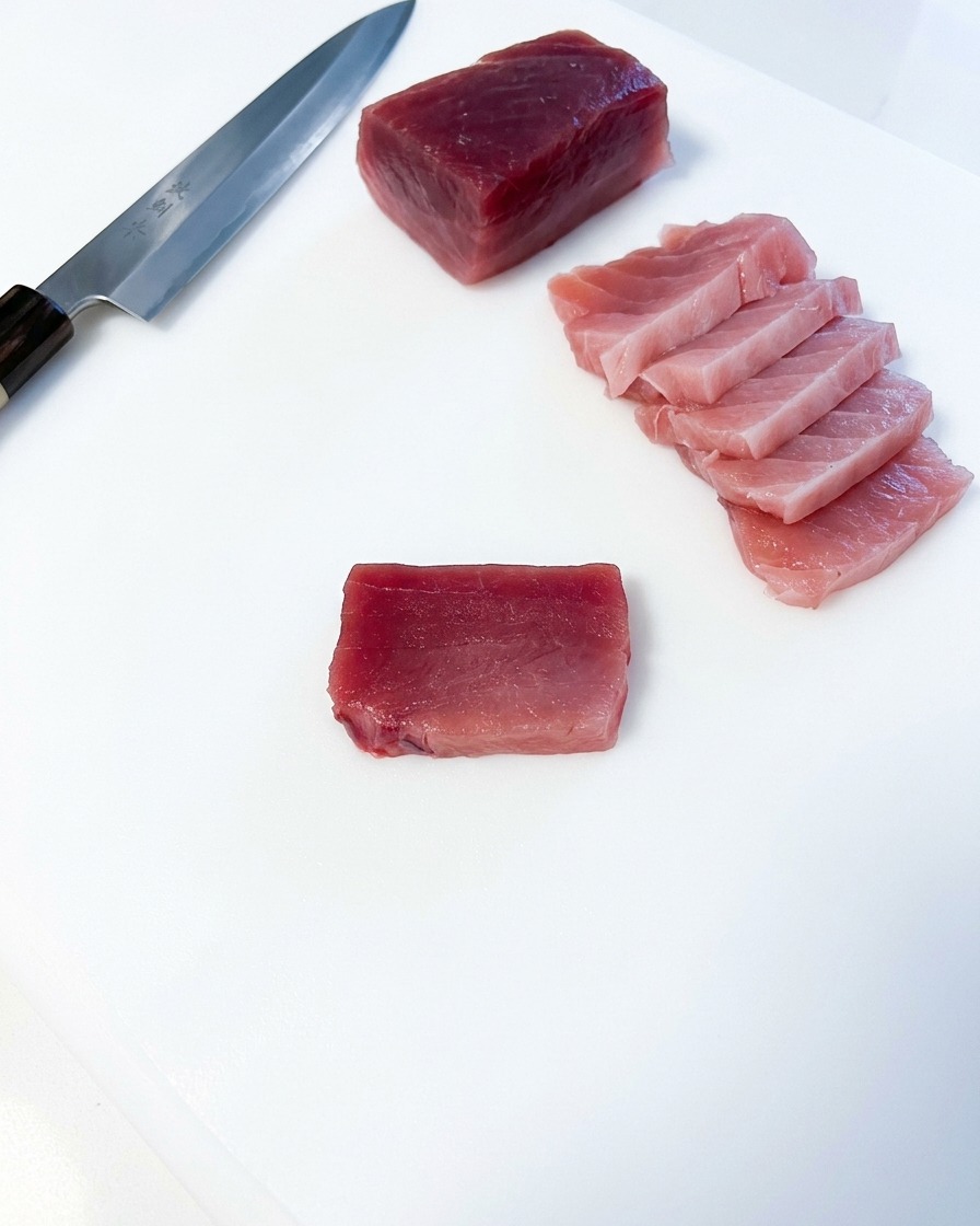 Varias rodajas rectangulares de atún graso rosado dispuestas junto a un gran bloque de atún y un cuchillo de cocina japonés sobre una tabla blanca.