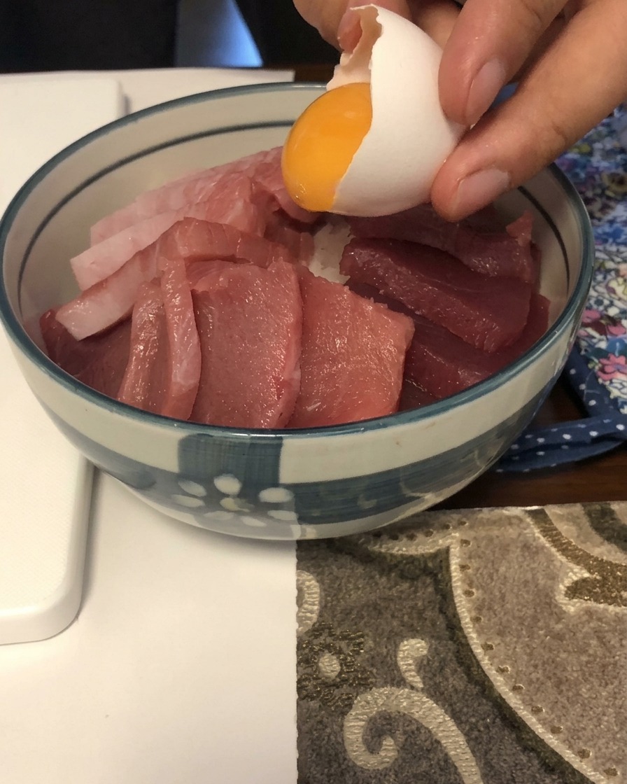 Una yema de huevo amarilla brillante deslizándose desde una cáscara blanca sobre una cama de pescado crudo en rodajas en un bol azul y blanco.