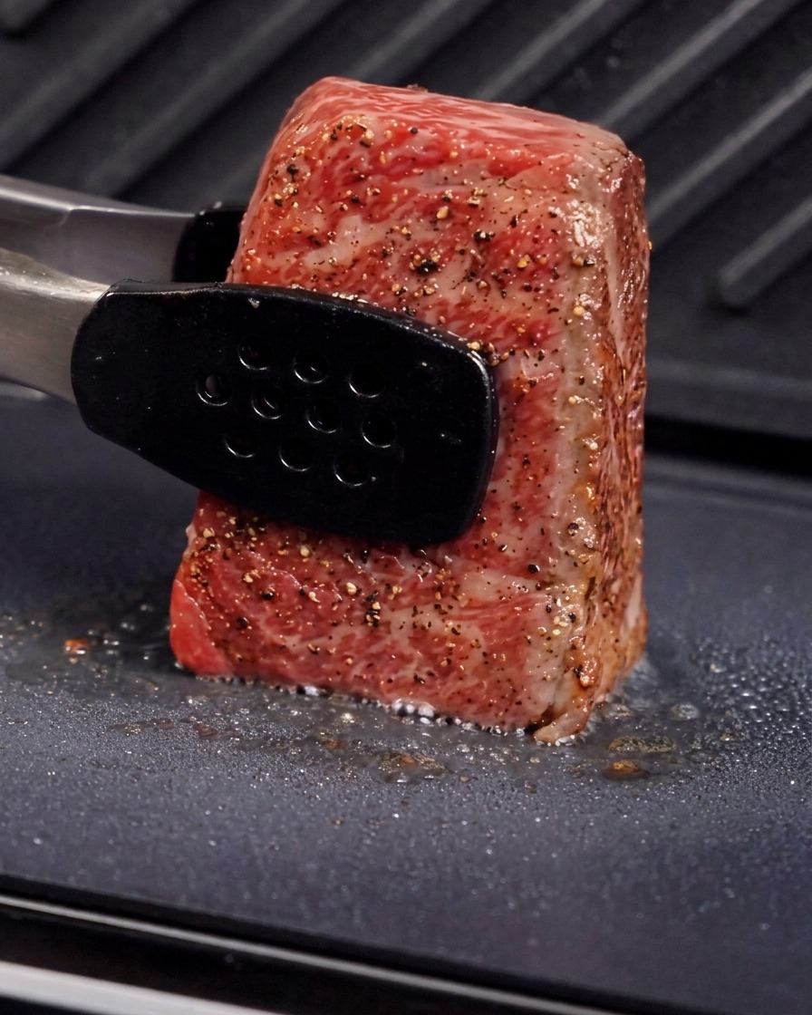 Pinzas de metal que sostienen un trozo grueso de carne en posición vertical sobre la placa de una parrilla para sellar el lateral, con vapor visible y aceite chisporroteando.
