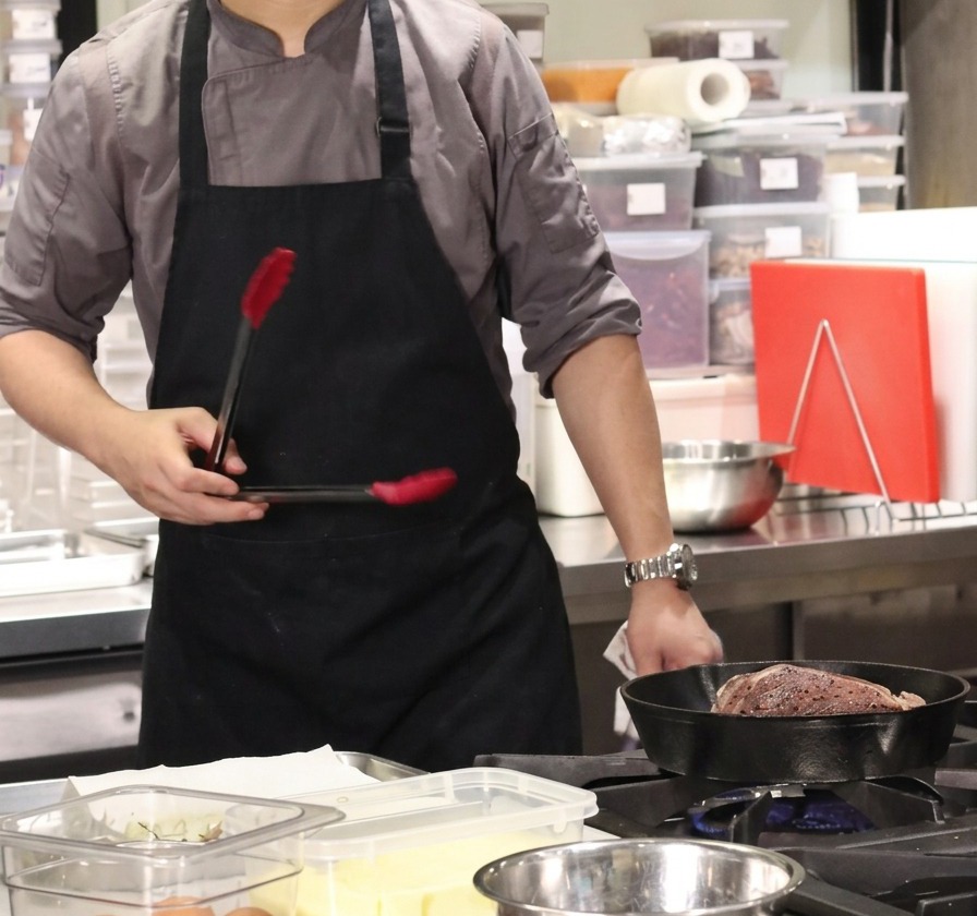 Un chef sosteniendo unas pinzas sobre una sartén de hierro fundido caliente con un gran bloque de ternera para sellar la capa de grasa.