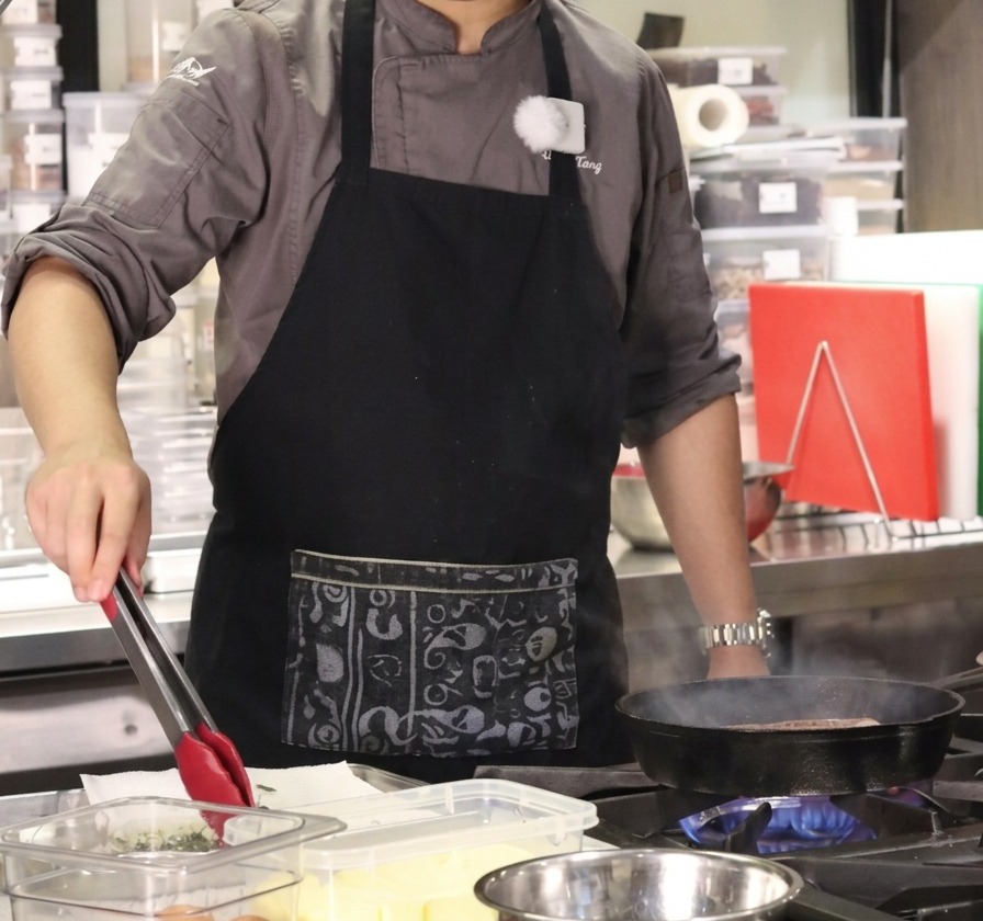 El chef usando pinzas para pasar ramitas de tomillo fresco y dientes de ajo desde un recipiente transparente a la sartén.