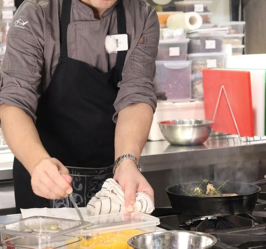 Un chef usando una cuchara para añadir un cubo de mantequilla desde un recipiente a una sartén humeante con carne y hierbas.