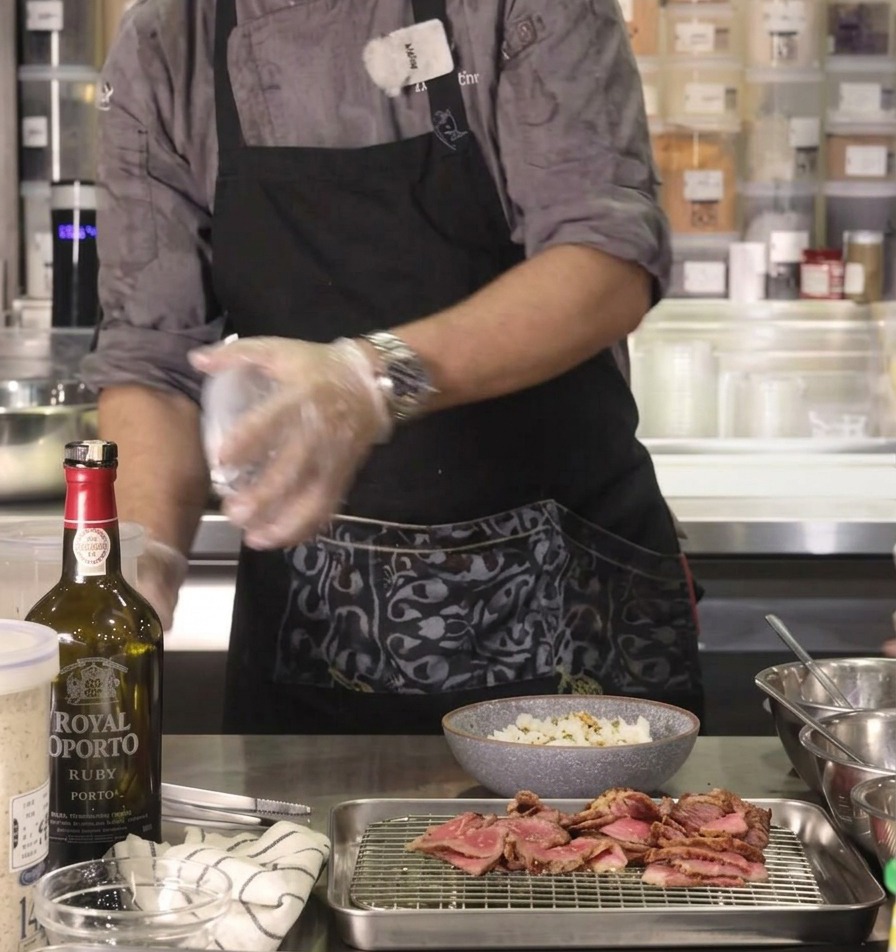 La mano del chef espolvoreando sazonador furikake sabroso sobre un bol de arroz en una encimera de cocina de acero inoxidable.