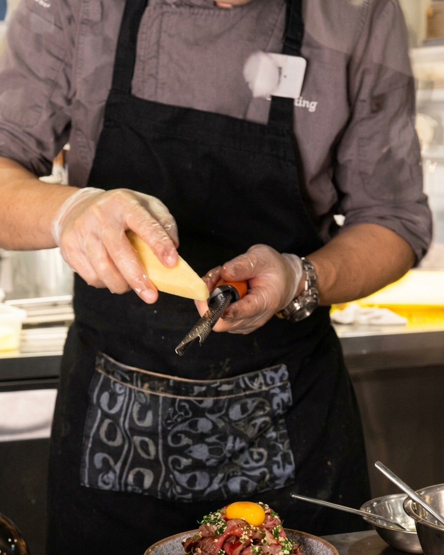 Un primer plano de un chef usando un rallador fino para rallar queso delicadamente sobre una imponente montaña de carne laminada coronada con una yema de huevo.