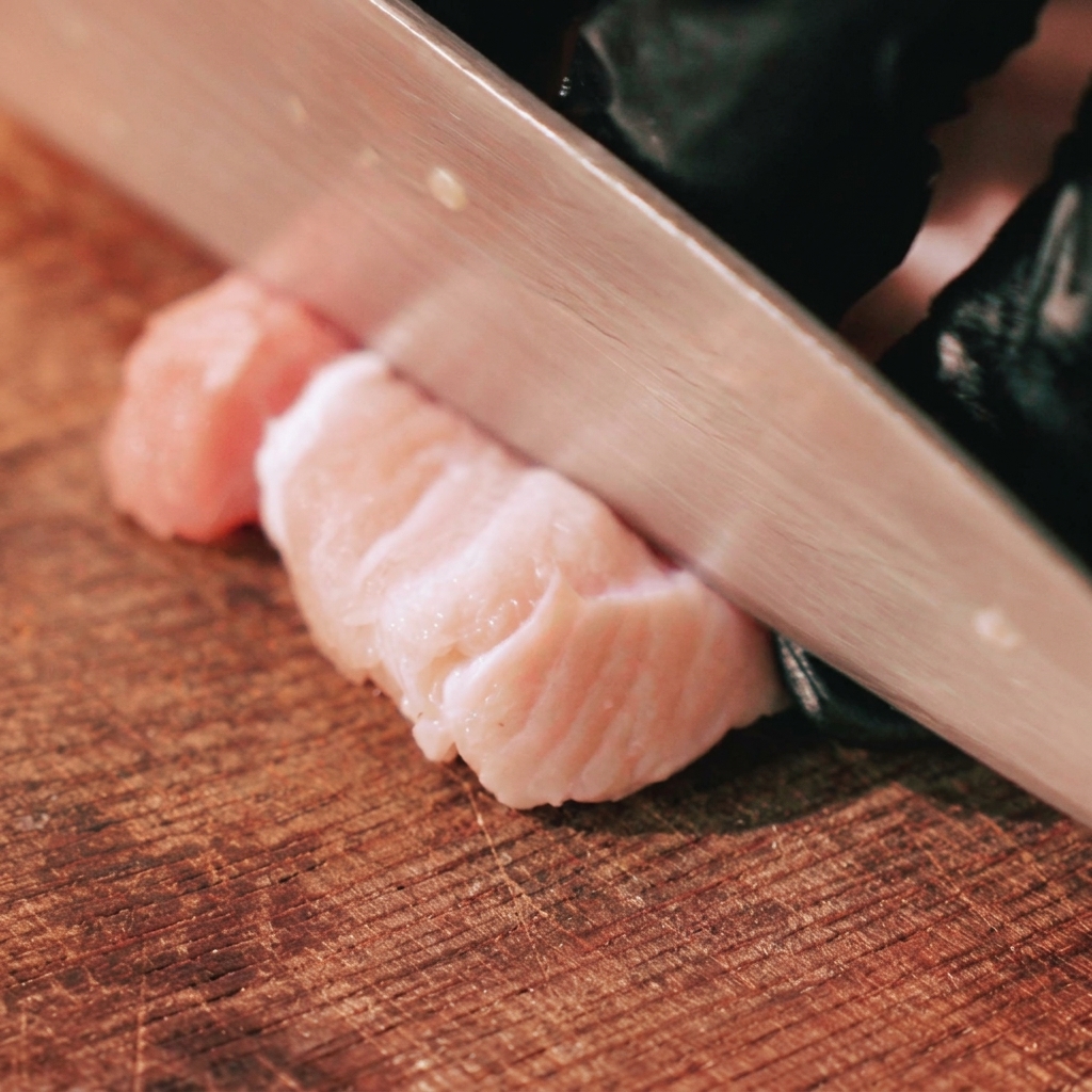 Una mano con guante usando un cuchillo de chef para cortar panceta de cerdo cruda con piel en tiras gruesas sobre una tabla de madera.