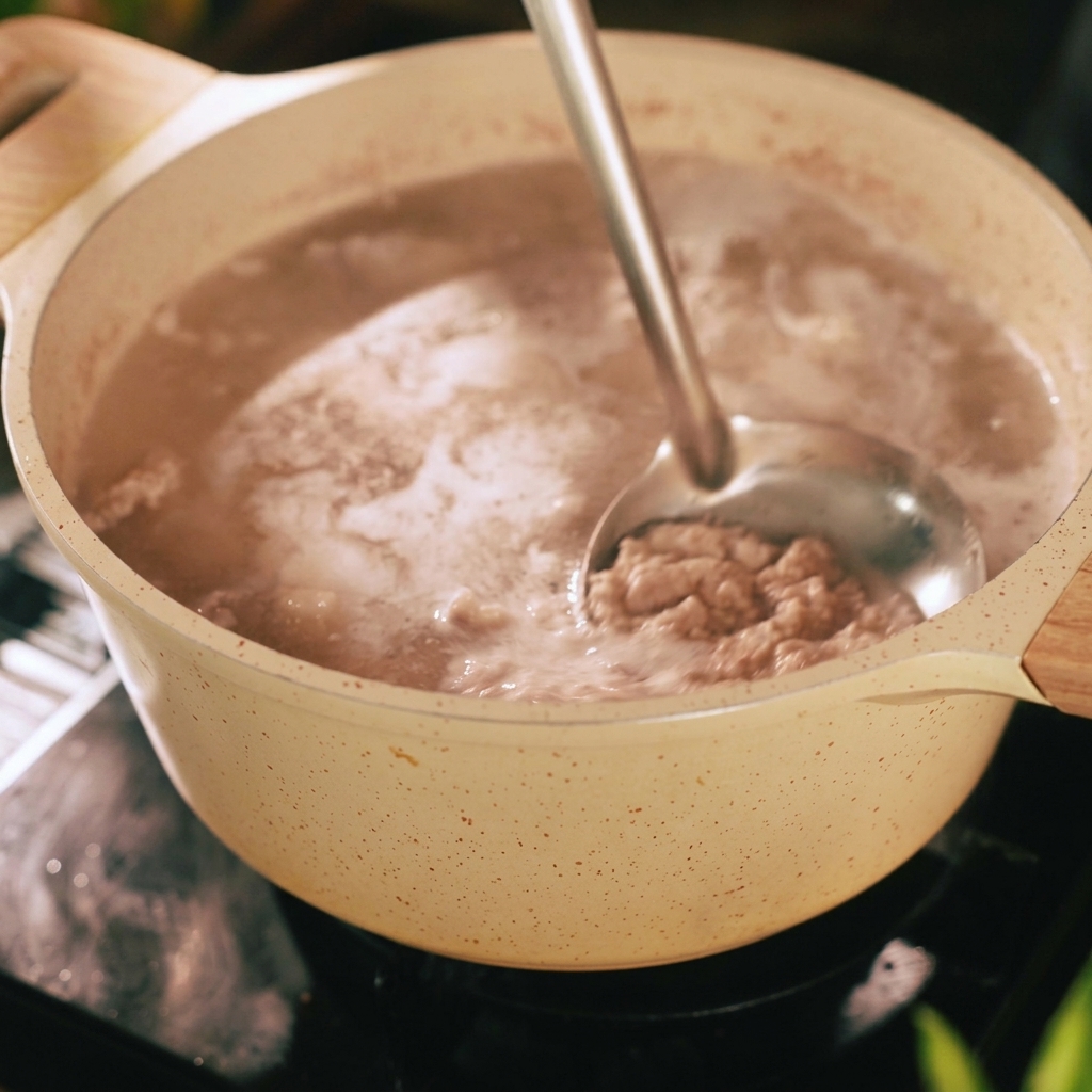 Un cazo de metal retirando espuma gris e impurezas de la superficie de agua hirviendo en una olla beige que contiene tiras de cerdo.
