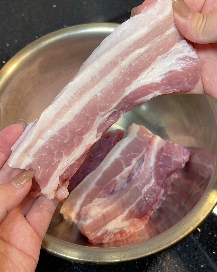 Dos manos sosteniendo una tira larga de panceta de cerdo cruda con capas rosadas y blancas, sin la piel, sobre un tazón de acero inoxidable.