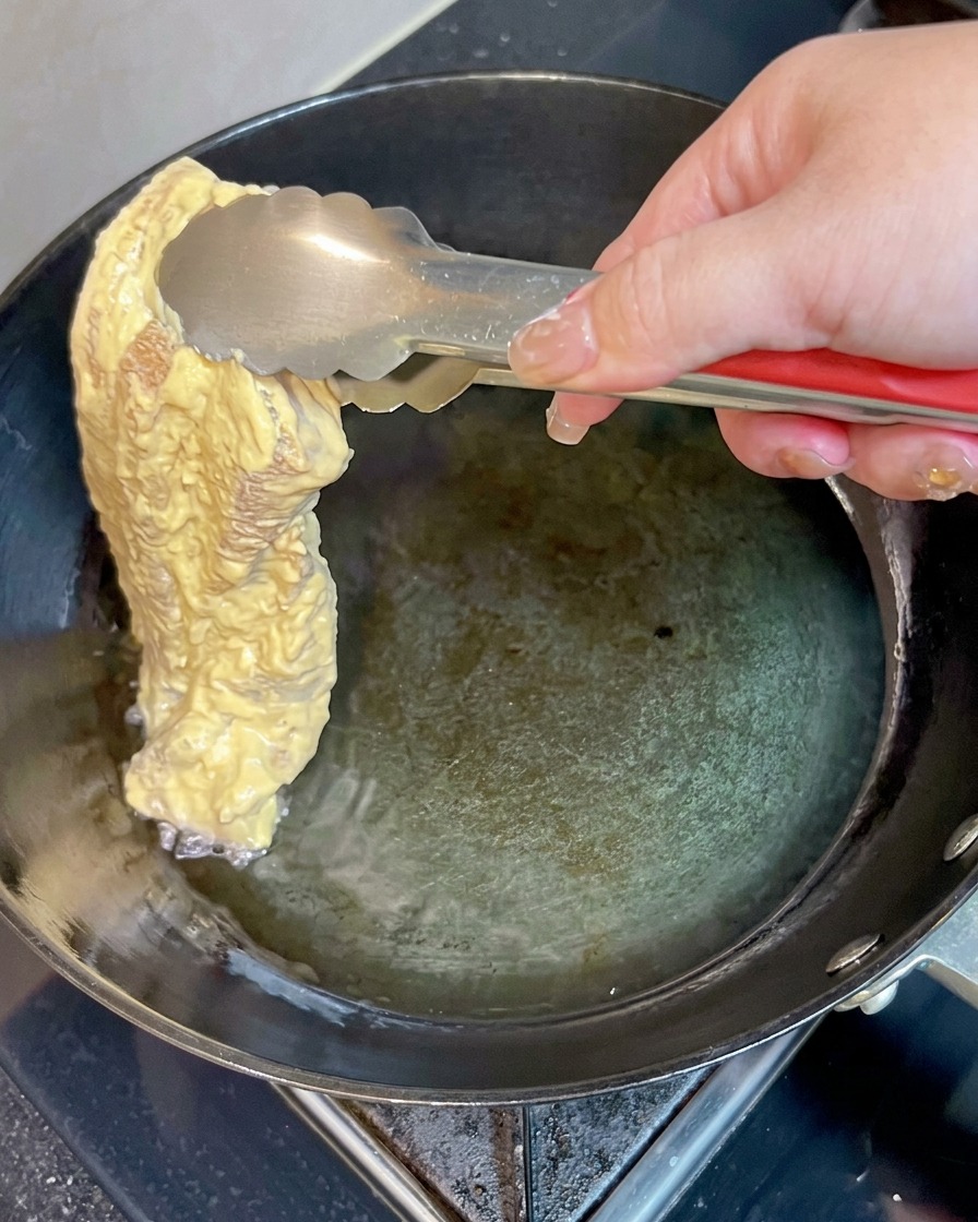 Un trozo de panceta de cerdo rebozada hundiéndose suavemente en aceite caliente dentro de una sartén usando pinzas de metal.