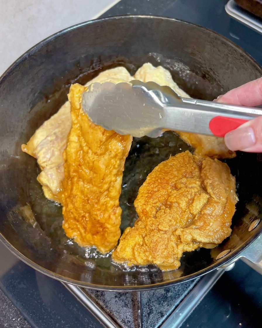 Pinzas de metal dando la vuelta a un trozo de panceta de cerdo crujiente y dorado en aceite caliente junto a otros trozos.