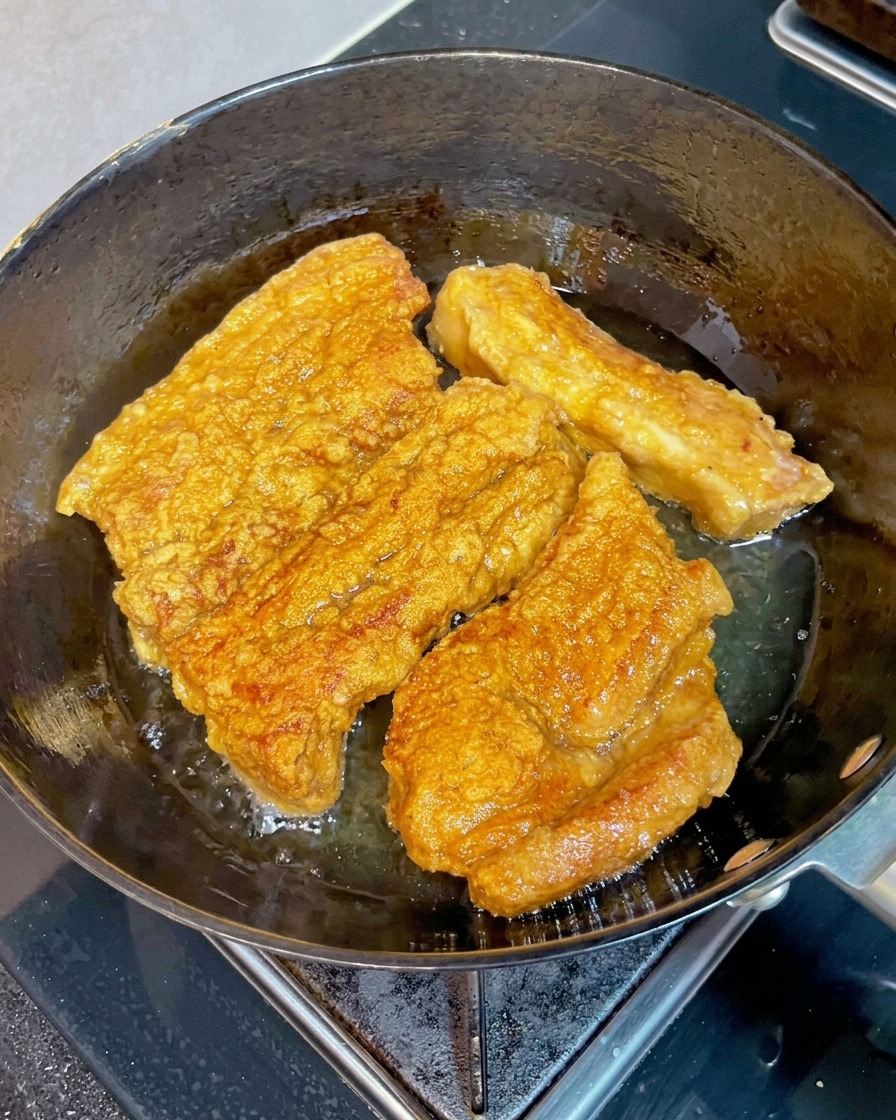 Tiras de panceta de cerdo doradas y crujientes friéndose en una sartén con aceite caliente mientras un palillo de madera presiona un trozo para comprobar su ternura.