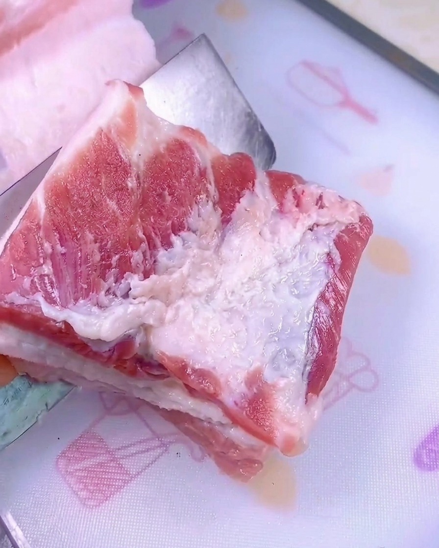 Un cuchillo afilado cortando un trozo grueso de panceta de cerdo cruda sobre una tabla de picar plástica blanca.