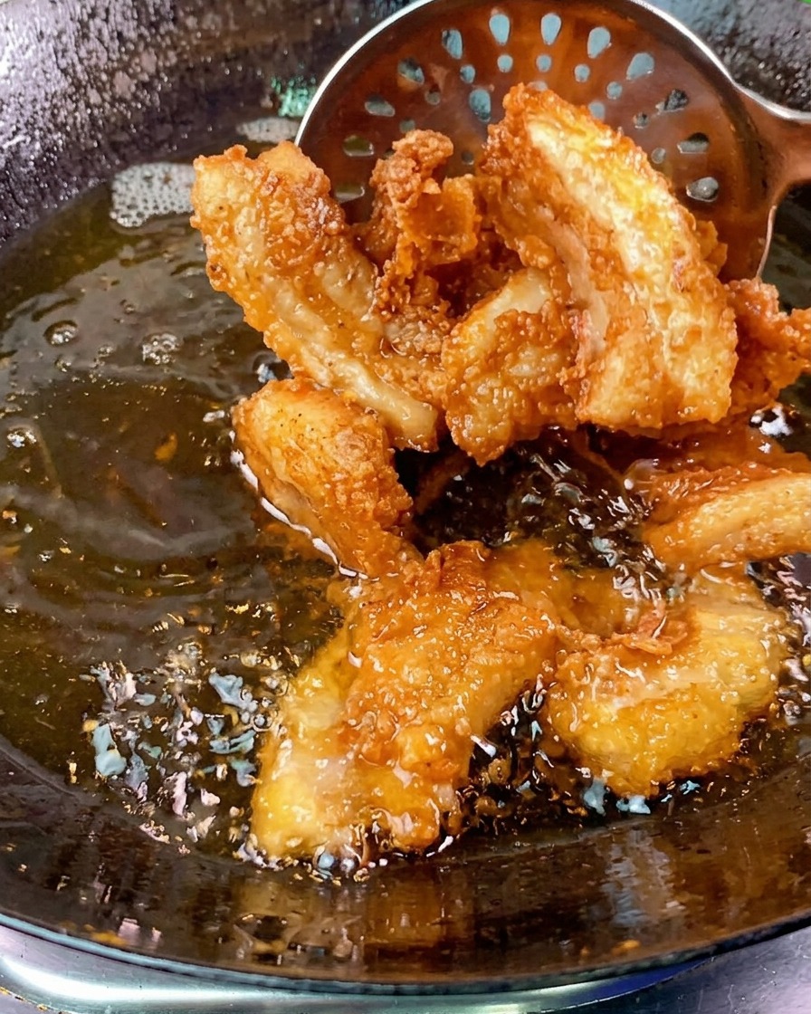 Trozos crujientes y dorados de panceta de cerdo siendo retirados de un wok con aceite burbujeante usando una espumadera metálica.