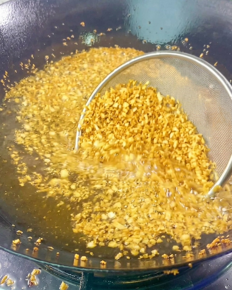Un colador de malla lleno de ajo picado sumergido en aceite caliente de fritura mientras se dora y suelta aroma.