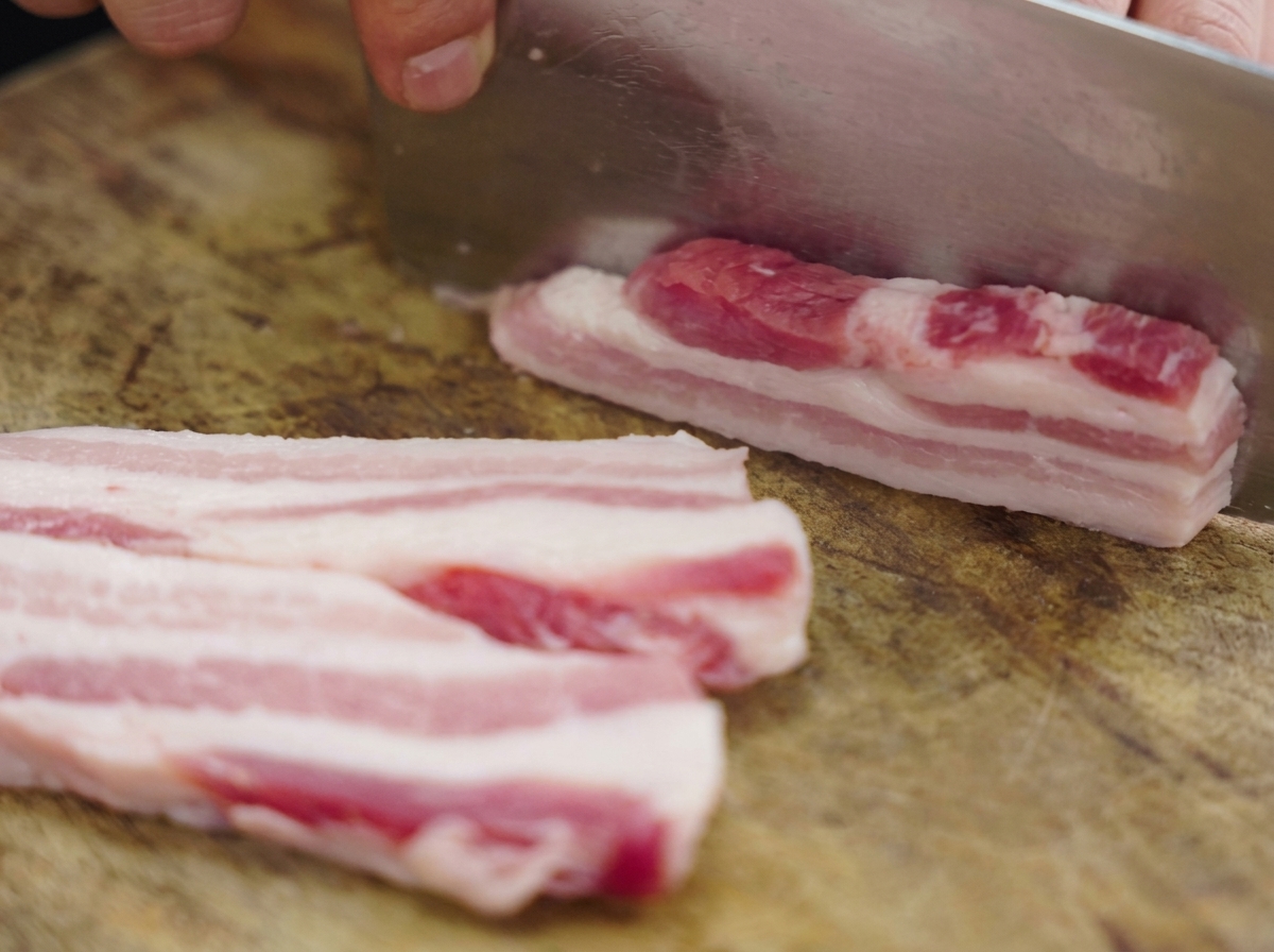 Tiras gruesas de panceta de cerdo cruda cortadas en una tabla de madera con un gran hacha de cocina metálica.