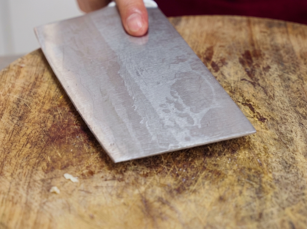 Un hacha de cocina de metal presionando plano sobre una tabla de cortar de madera para machacar dientes de ajo crudos.