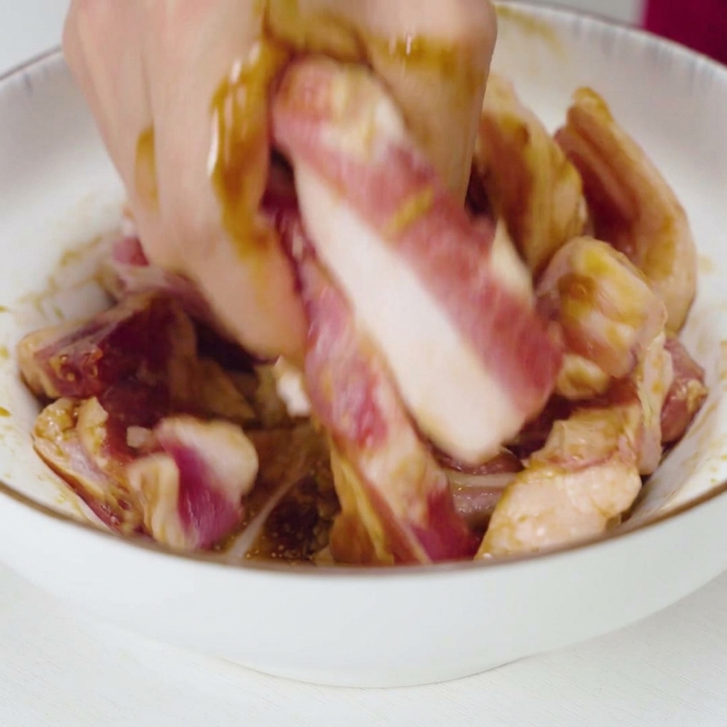 Una mano mezclando y masajeando tiras de cerdo crudo con un marinado líquido oscuro y trozos de ajo en un bol blanco.