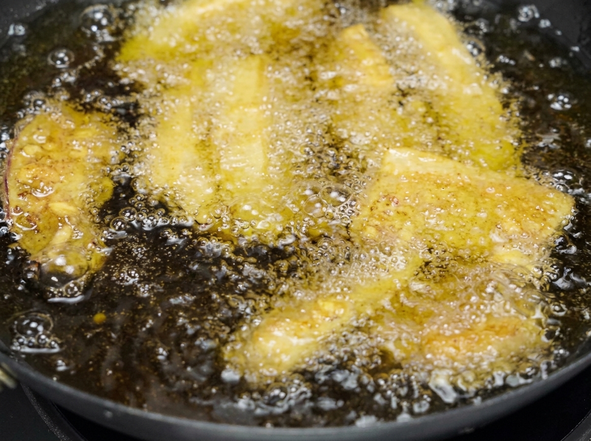 Varias tiras de panceta de cerdo rebozadas friéndose en una sartén llena de aceite dorado burbujeante.