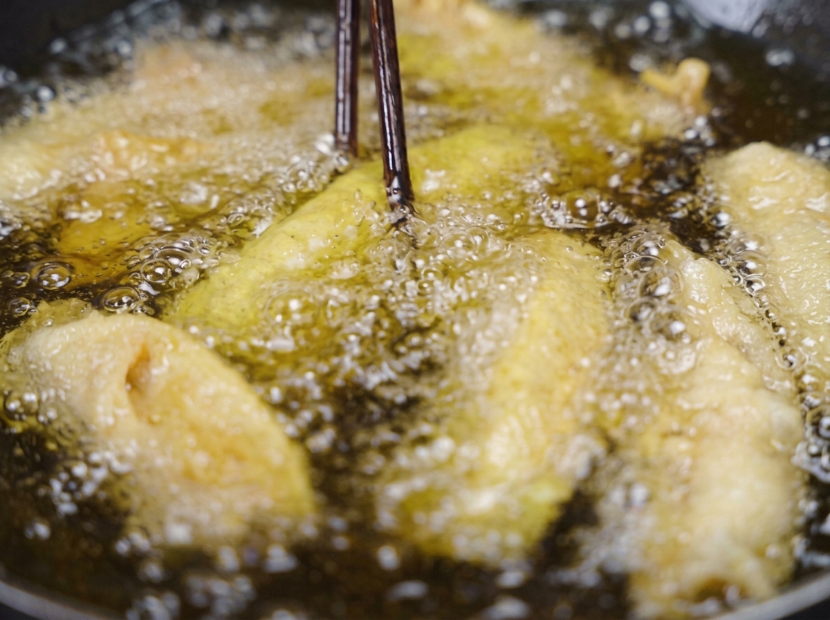 Unos palillos de madera oscura levantando una tira de cerdo frita, dorada y crujiente, del aceite de cocina burbujeante.