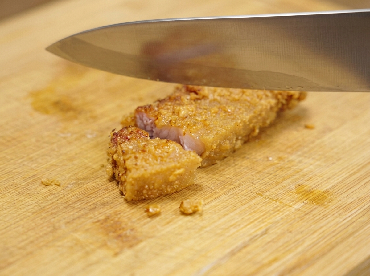 Un gran cuchillo de metal cortando un trozo de cerdo frito crujiente y dorado en porciones pequeñas sobre una tabla de cortar de madera.